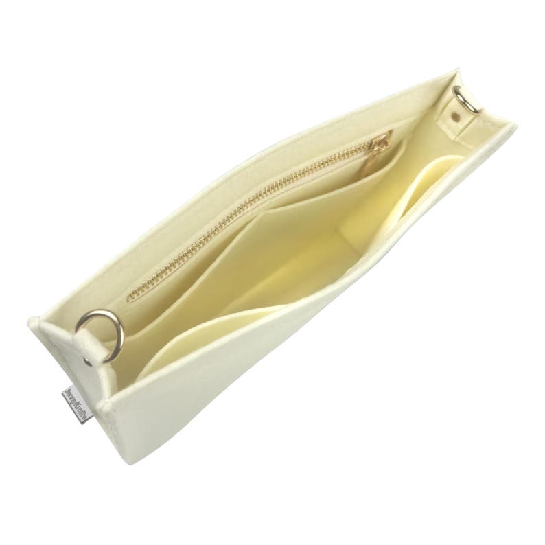 May include: Cream-coloured felt handbag organiser with multiple compartments and a zipped pocket. The interior is a lighter shade of cream. Features a gold-coloured zip and metal hardware for attaching a strap. The organiser is designed to fit inside a larger bag.