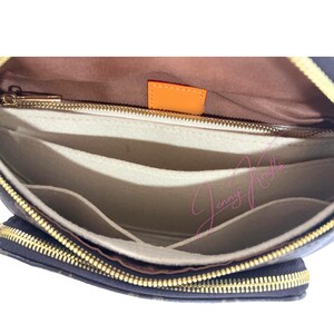 May include: Open view of a navy blue handbag interior with multiple compartments. The interior features a light tan lining and several felt dividers. A gold zipper and orange tag are visible.