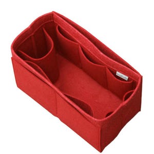 May include: A rectangular red felt handbag organizer insert featuring multiple interior compartments and pockets of varying sizes to help keep items organized. A small white rectangular tag is attached to the inner side wall of the felt structure.