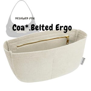 Può includere: Un organizer per borsa in feltro color bianco sporco con molteplici scomparti interni, una chiusura a zip in metallo dorato e una piccola etichetta laterale. Il testo "DESIGNED FOR Coa*.Belted Ergo" è stampato in nero sopra l'organizer.