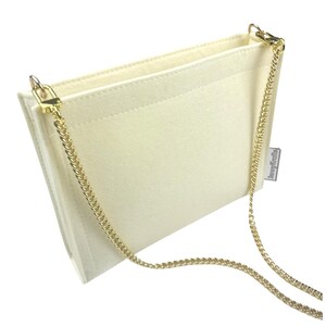 May include: Cream-coloured felt handbag with a gold chain strap. The bag has a rectangular shape with a flap closure and a gold chain strap. The interior is visible, and the bag appears to be empty. The bag has a small tag that says "JennyKrafts".