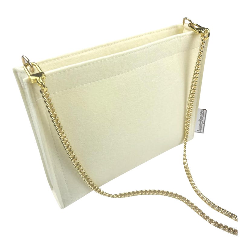 May include: Cream-coloured felt handbag with a gold chain strap. The bag has a rectangular shape with a flap closure and a gold chain strap. The interior is visible, and the bag appears to be empty. The bag has a small tag that says "JennyKrafts".