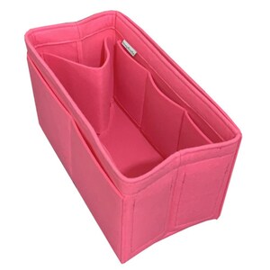 May include: A bright pink felt handbag organizer insert with a rectangular shape. The interior features multiple vertical dividers and open compartments for storage. A small white fabric tag with black text is attached to the inner side wall.