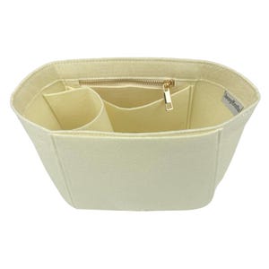Op de afbeelding: Cr&egrave;mekleurige vilten handtassenorganizer met meerdere compartimenten en een vak met ritssluiting. De binnenkant heeft verschillende vakken voor organisatie. Een goudkleurige rits en trekker zijn zichtbaar. De organizer is ontworpen om in een grotere tas te passen.