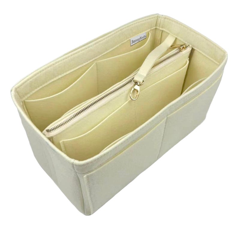 May include: Cream-coloured felt handbag organiser with multiple compartments and a zipped pouch. The organiser has a rectangular shape with several pockets and a zipped pouch with a cream-coloured strap and gold-coloured hardware. The brand name is visible on the top.