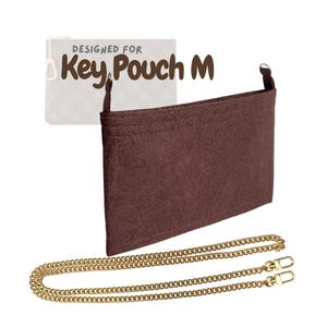 Puede incluir: Un organizador de fieltro marrón con el texto "DESIGNED FOR Key Pouch M" y una cadena dorada. El organizador tiene múltiples compartimentos y está diseñado para caber dentro de un llavero. La cadena tiene dos cierres.