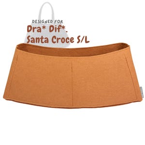 Peut inclure: Un organisateur de sac en feutre beige avec le texte "DESIGNED FOR Dra* Dif*. Santa Croce S/L". L'organisateur a un dessus incurvé et une forme rectangulaire avec des coutures verticales. Le fond est blanc.