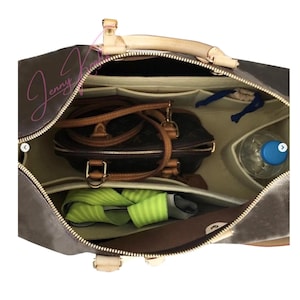 Puede incluir: Vista interior de un bolso marrón con múltiples compartimentos. En su interior, se observa un bolso marrón más pequeño, una correa verde y blanca, una botella de agua y otros objetos. El bolso tiene un asa y ribetes beige.