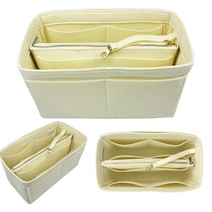 May include: Cream-coloured felt handbag organiser with multiple compartments and a zipped pouch. The organiser has a rectangular shape with a structured design, featuring a handle and a gold-toned zip.