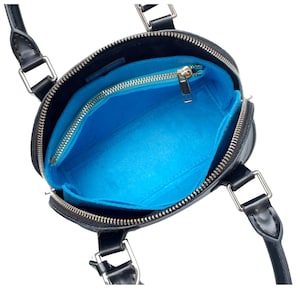 Puede incluir: Bolso de mano negro y redondo con herrajes plateados y cremallera superior. El interior presenta un organizador de fieltro azul brillante con cremallera dorada. El bolso tiene dos asas cortas y una bandolera.
