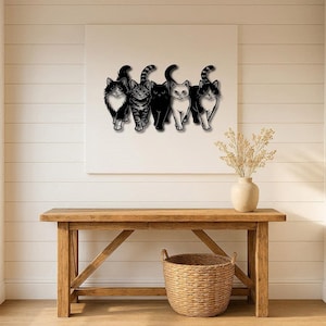 May include: A black metal wall art piece featuring five stylized cats in various poses. The cats are in black with white accents, and one has a striped tail. The artwork is mounted on a white canvas above a wooden table.