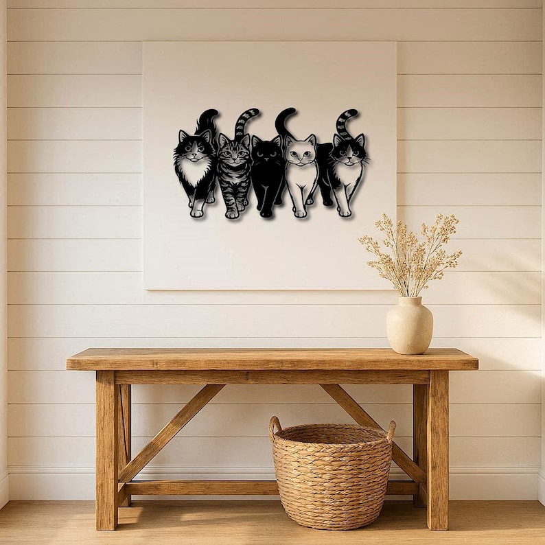 May include: A black metal wall art piece featuring five stylized cats in various poses. The cats are in black with white accents, and one has a striped tail. The artwork is mounted on a white canvas above a wooden table.