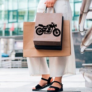 May include: A person holding two shopping bags. The front bag is brown with a black motorcycle graphic. The second bag is also brown. The person is wearing black sandals and white pants. The background is a modern building.