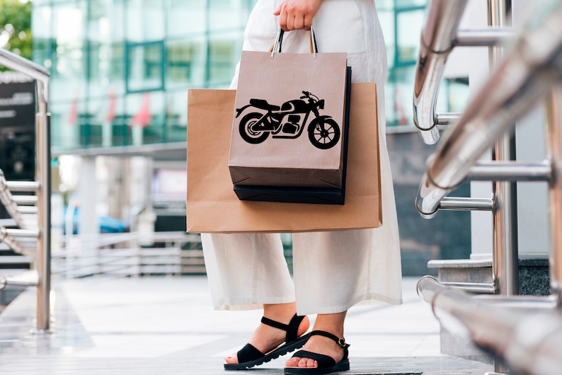 May include: A person holding two shopping bags. The front bag is brown with a black motorcycle graphic. The second bag is also brown. The person is wearing black sandals and white pants. The background is a modern building.