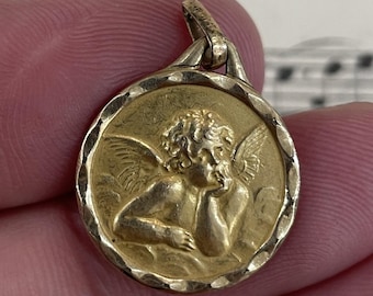 May include: A gold-colored pendant featuring a detailed relief of a cherub with wings, resting its head on its hand. The pendant has a textured, circular border and a small loop for a chain.