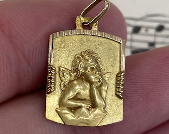 May include: A gold-colored pendant with a rectangular shape and a small loop at the top. The pendant features a detailed relief of a cherub with wings, resting its head on its hand. The cherub is set against a textured background.