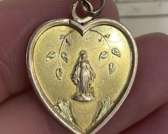 May include: Gold-toned heart-shaped pendant with a detailed religious figure in the center. The pendant features a gold border and a textured gold background with engraved leaf and vine details. A small loop is at the top for a chain.