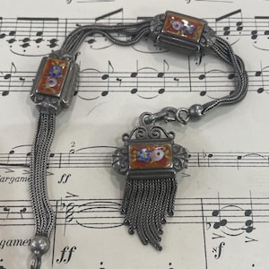 Puede incluir: Un brazalete de cadena de plata con dos dijes de esmalte con diseños florales en tonos rojos, azules y blancos. El brazalete tiene un colgante de borla.