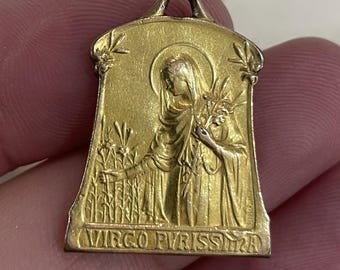 May include: A gold-colored pendant featuring a religious figure with a halo, holding a sprig of leaves. The pendant has the words "VIRGO PURISSIMA" at the bottom and is rectangular with a curved top.