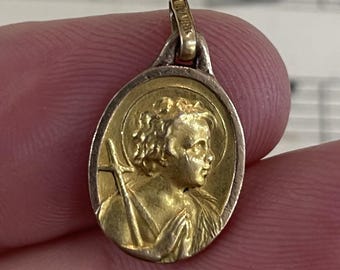 May include: Gold-toned pendant with an oval shape, featuring a detailed relief of a figure with a halo and a cross. The pendant has a loop at the top for a chain. The metal has a vintage appearance.