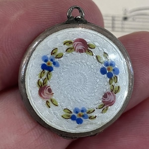 May include: A round, silver-toned pendant with a white enamel face. The face is decorated with a wreath of pink roses, blue forget-me-nots, and green leaves. The pendant has a small loop at the top for a chain.