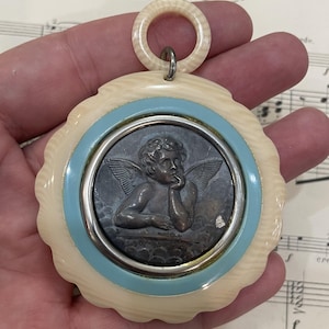 May include: A vintage pendant featuring a cherub relief within a silver-toned frame, surrounded by a light blue and cream-colored scalloped border. The pendant has a cream-colored ring at the top for hanging.