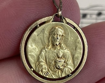 May include: A gold-colored pendant featuring a detailed relief of a religious figure holding a heart. The pendant is circular with a loop at the top for a chain. The design is set within a decorative frame.
