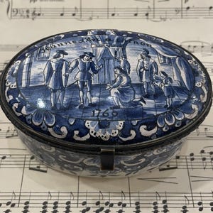 Op de afbeelding: Een ovale, blauw-witte keramische doos met een scharnierend deksel. Het deksel toont een scène met mensen in een kamer met de datum 1768. De doos is versierd met een bloemmotief.