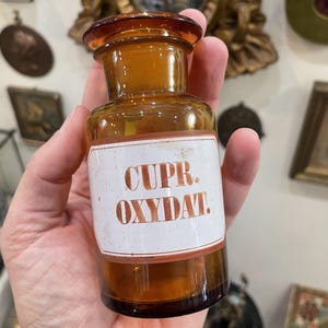 Może przedstawiać: Zabytkowa butelka z brązowego szkła z brązowym korkiem. Butelka ma białą etykietę z napisem "CUPR. OXYDAT." w brązowych literach. Butelka jest trzymana w dłoni.