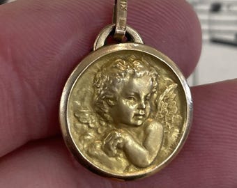 May include: A gold-colored pendant with a detailed relief of a cherub. The cherub has curly hair and is depicted with wings. The pendant has a loop at the top for a chain and the marking "XII" is visible.