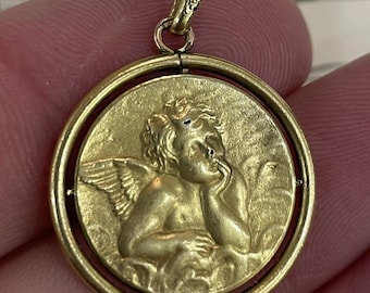 May include: A gold-colored pendant featuring a detailed relief of a cherub with wings, resting its chin on its hand. The cherub is set within a circular frame. The pendant is suspended from a small loop at the top.
