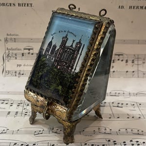 Puede incluir: Marco de fotos antiguo de cristal y latón con una imagen pintada de una catedral en una colina, con la inscripción "Lyon. N.D. de Fourviere". El marco tiene detalles ornamentales de latón y un soporte con bisagras. Fondo de partituras.