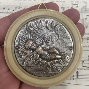 May include: A round, cream-colored, plastic frame with a silver, embossed image of a baby Jesus lying in a manger. The image is surrounded by a halo of light and clouds.