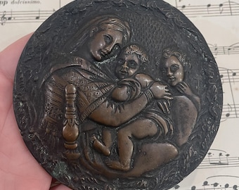 May include: A bronze circular plaque depicting a mother and two children. The mother is holding a child in her arms, while the other child is standing next to her. The plaque is decorated with a floral design around the edge.