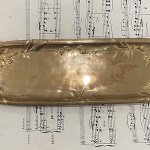 Op de afbeelding: Een vintage, rechthoekige messing dienblad met sierlijke bloemen- en bladmotieven. De lade heeft een licht gebogen vorm met opstaande randen en ligt op een vel antieke bladmuziek. Het oppervlak van de lade heeft een warme, gouden tint.