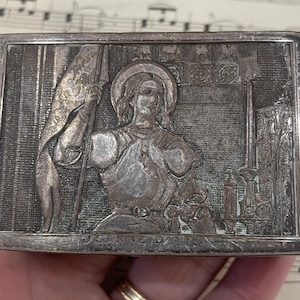 Op de afbeelding: Een rechthoekige, zilverkleurige doos met een reliëfbeeld van Jeanne d'Arc met een vlag. De afbeelding bevat architecturale details en de tekst "JEANNE D'ARC" onderaan. De doos heeft een licht aangetaste uitstraling.