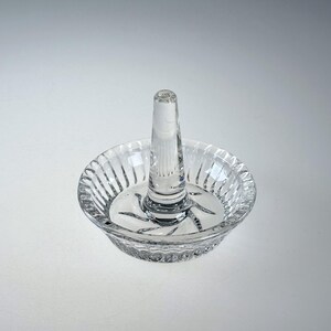 May include: A clear glass ring holder, featuring a central post and a shallow, circular base. The base has a ribbed design around the edge and a starburst pattern in the centre. Designed to hold rings and other small jewellery items.