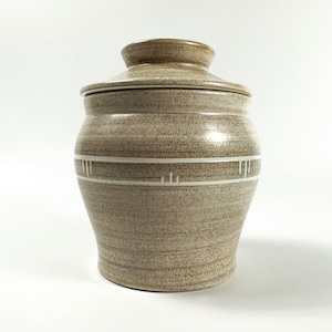 May include: A beige ceramic jar with a lid, featuring a white band with a geometric design around the middle.