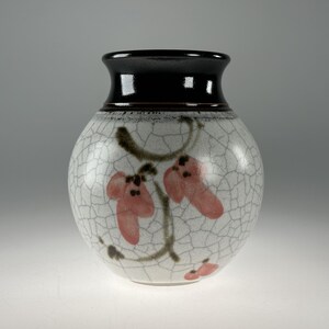 Könnte beinhalten: Kleine, runde Keramikvase mit schwarzem Rand und weißem, craquelé Körper. Die Vase ist mit rosa Blumendesigns und braunen Stielen verziert. Die Vase ist ein dekorativer Gegenstand.