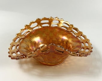 May include: An orange iridescent glass basket with a woven design and a decorative, openwork rim. The basket has a handle on each side. The glass has a shimmering, metallic finish. The basket is designed to hold small items.