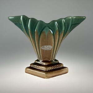 May include: A ceramic vase with a fan-shaped top and a square base. The vase has a two-tone colour scheme, with a green top and a tan body. The base is a darker shade of tan. A small, oval-shaped label is on the front.