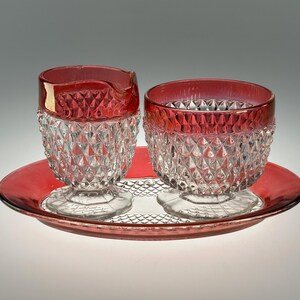 Könnte beinhalten: Ein Vintage-Set aus rotem Glas mit Sahnekännchen und Zuckerdose mit Rautenmuster, das auf einem passenden ovalen Tablett steht. Die Ränder des Sahnekännchens und der Schüssel sind tieferrot.