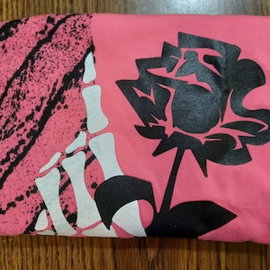 Peut inclure: Un t-shirt rose avec une empreinte de main de squelette noir et blanc sur le c&ocirc;t&eacute; gauche et une rose noire sur le c&ocirc;t&eacute; droit.
