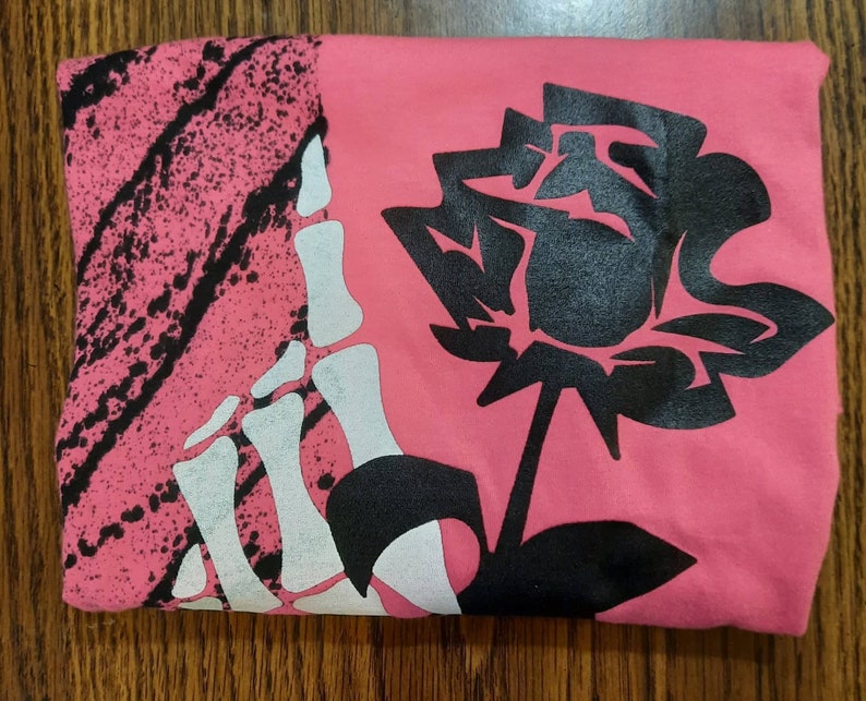 Peut inclure: Un t-shirt rose avec une empreinte de main de squelette noir et blanc sur le c&ocirc;t&eacute; gauche et une rose noire sur le c&ocirc;t&eacute; droit.