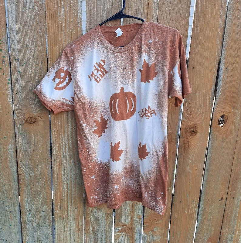 May include: A brown t-shirt with a bleach-splattered design featuring a pumpkin, maple leaves, and the text "Hello Fall" and "Farm Fresh".