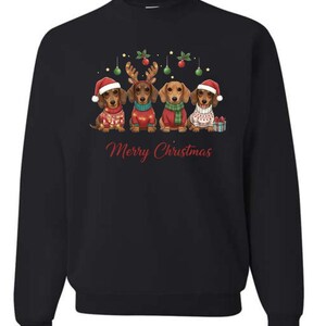 May include: Black sweatshirt featuring a festive design with four dachshunds dressed in holiday attire. Each dog wears a Santa hat, reindeer antlers, or a Christmas sweater. The words "Merry Christmas" are written in red script.