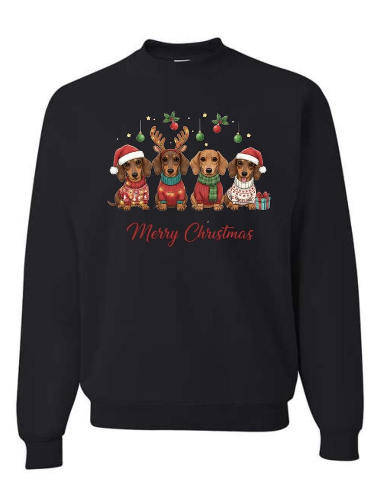 May include: Black sweatshirt featuring a festive design with four dachshunds dressed in holiday attire. Each dog wears a Santa hat, reindeer antlers, or a Christmas sweater. The words "Merry Christmas" are written in red script.