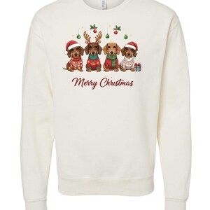 May include: Cream-colored sweatshirt featuring a festive design with four dachshunds dressed in holiday attire. Each dog wears a Santa hat, reindeer antlers, or a sweater. The words "Merry Christmas" are written in a cursive font.