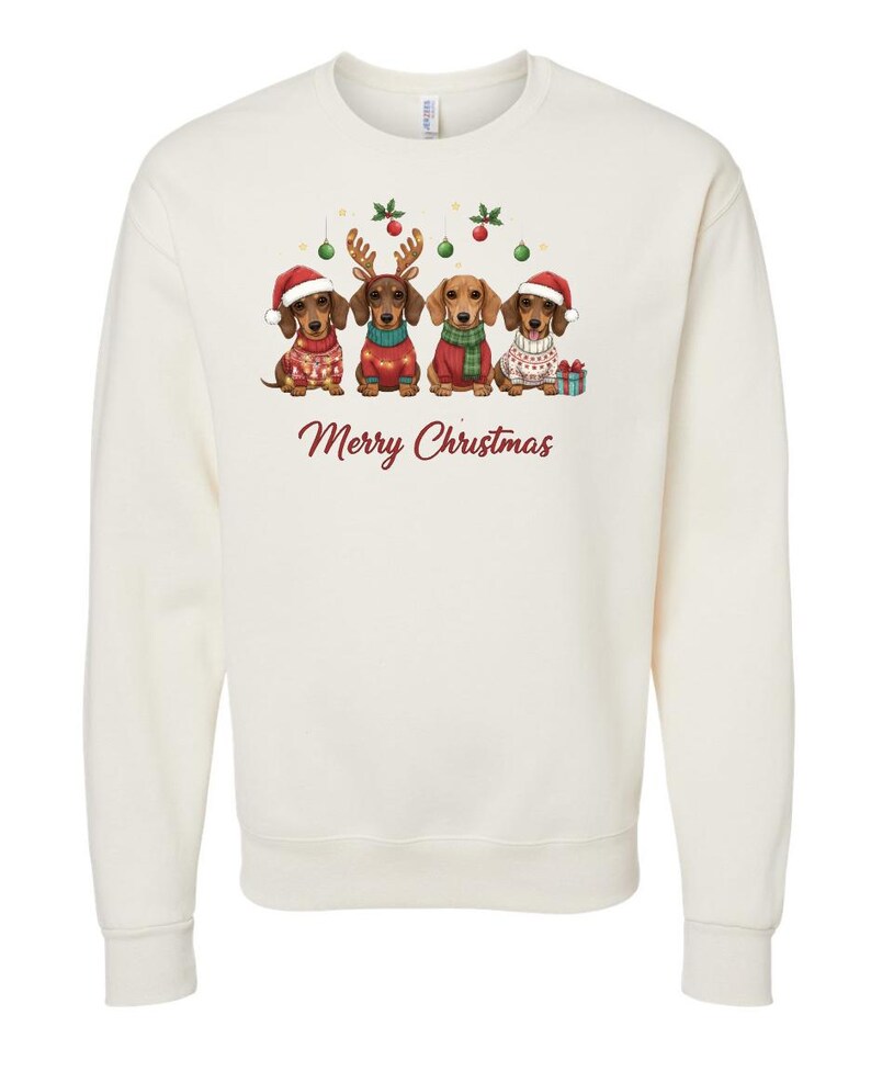 May include: Cream-colored sweatshirt featuring a festive design with four dachshunds dressed in holiday attire. Each dog wears a Santa hat, reindeer antlers, or a sweater. The words "Merry Christmas" are written in a cursive font.