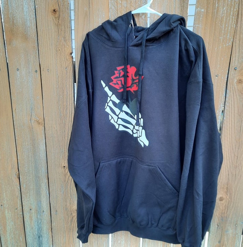 Peut inclure: Sweat-shirt &agrave; capuche noir avec un graphique de main squelettique blanche tenant une rose rouge.